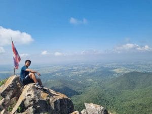 A man sat on top of a mountain, showing grassland and hills beyond and a Thailand flat is being flown behind him.