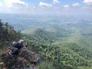A man sat on top of a mountain, showing grassland and hills beyond.