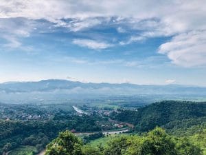 A wide scape view of a town in Thailand, with green trees in the foreground and a cloudy but blue sky.