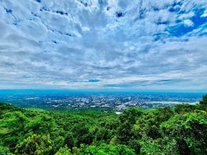 A wide scape view of a town in Thailand, with green trees in the foreground and a cloudy but blue sky.